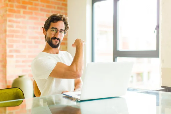 young bearded man with a laptop feeling happy, satisfied and powerful, flexing fit and muscular biceps, looking strong after the gym