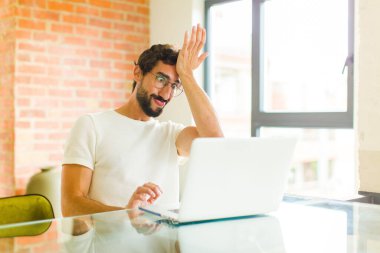 young bearded man with a laptop raising palm to forehead thinking oops, after making a stupid mistake or remembering, feeling dumb