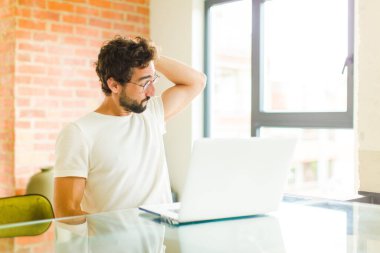 Dizüstü bilgisayarı olan genç sakallı bir adam bilgisiz ve kafası karışık, bir çözüm düşünüyor, eli kalçasında ve diğer kafasında, dikiz aynasından