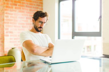 Dizüstü bilgisayarı olan sakallı genç adam hayal kırıklığına uğramış ve hayal kırıklığına uğramış görünüyor. Ciddi görünüyor, kızgın ve kollarını kavuşturmuş.