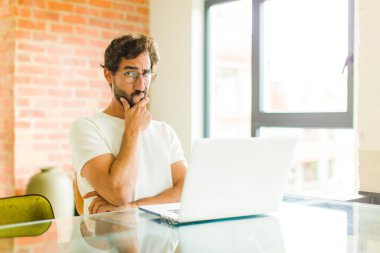 Dizüstü bilgisayarı olan genç sakallı bir adam ciddi, düşünceli ve güvensiz görünüyor, bir kolu çapraz ve çenesi üzerinde, seçenekleri tartıyor.