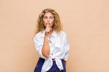 Orta yaşlı kadın ciddi görünüyor ve parmaklarını dudaklarına bastırıp sessizlik ya da sessizlik talep ediyor, sır saklıyor.