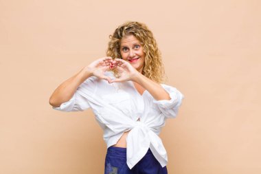Orta yaşlı bir kadın gülümsüyor ve mutlu, sevimli, romantik ve aşık, iki eliyle kalbi şekillendiriyor.