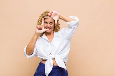 Orta yaşlı bir kadın mutlu, arkadaş canlısı ve pozitif hissediyor, gülümsüyor ve elleriyle bir portre veya fotoğraf çerçevesi yapıyor.