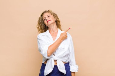 Orta yaşlı kadın neşeli bir şekilde gülümsüyor, mutlu hissediyor ve yan ve yukarı doğru işaret ediyor, fotokopi uzayındaki nesneleri gösteriyor.