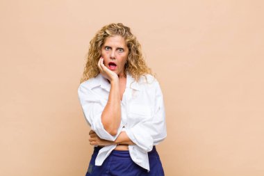 Orta yaşlı bir kadın şok ve inançsızlık içinde, elleri yanağında ve kolları çaprazlanmış, sersemlemiş ve şaşırmış hissediyor.