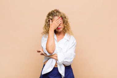 Orta yaşlı bir kadın stresli, utanmış ya da üzgün görünüyor, baş ağrısı var, yüzünü elle kapatıyor.