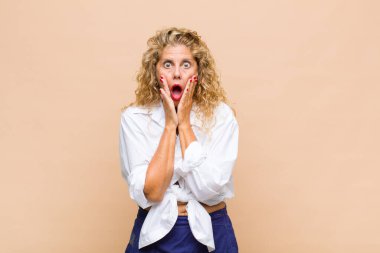 Orta yaşlı kadın şok olmuş ve korkmuş hissediyor, ağzı açık ve elleri yanaklarında korkmuş görünüyor.