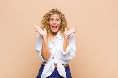 Orta yaşlı bir kadın mutlu ve heyecanlı görünüyor. Beklenmedik bir sürprizle şoke olmuş. İki eli yüzü açık.