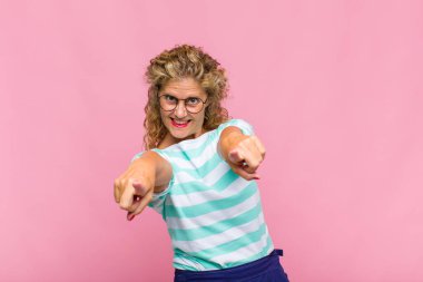 Orta yaşlı bir kadın mutlu ve kendinden emin, kamerayı iki eliyle gösteriyor ve gülüyor, seni seçiyor.