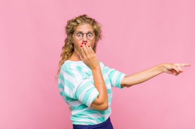 Orta yaşlı kadın kendini mutlu, şaşkın ve şaşırmış hissediyor. Ağzını eliyle kapatıyor ve yanal fotokopi alanını işaret ediyor.