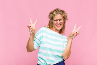 Orta yaşlı kadın gülümsüyor ve mutlu görünüyor, arkadaş canlısı ve memnun, zafer ya da barış işareti yapıyor iki eliyle