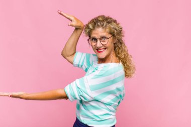 Orta yaşlı kadın gülümsüyor, mutlu, pozitif ve tatmin olmuş hissediyor, fotokopi alanında nesne veya konsept tutuyor veya gösteriyor