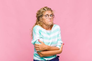 Orta yaşlı bir kadın omuz silkiyor, kafası karışık ve kararsız, kolları bağlı ve şaşkın görünüyor.