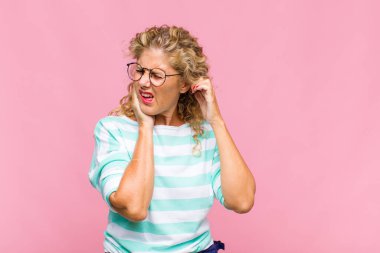 Orta yaşlı bir kadın sinirli, stresli ve sinirli görünüyor, iki kulağını da sağır edici bir ses, ses ya da yüksek sesle kapatıyor.