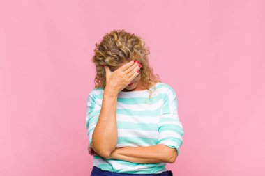 Orta yaşlı bir kadın stresli, utanmış ya da üzgün görünüyor, baş ağrısı var, yüzünü elle kapatıyor.