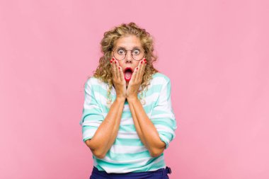 Orta yaşlı kadın şok olmuş ve korkmuş hissediyor, ağzı açık ve elleri yanaklarında korkmuş görünüyor.