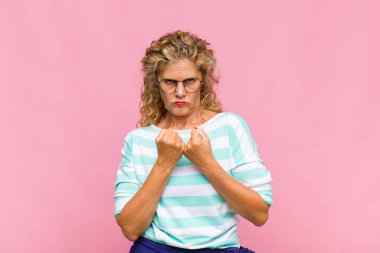 Orta yaşlı kadın kendinden emin, sinirli, güçlü ve agresif görünüyor. Yumrukları boks pozisyonunda dövüşmeye hazır.