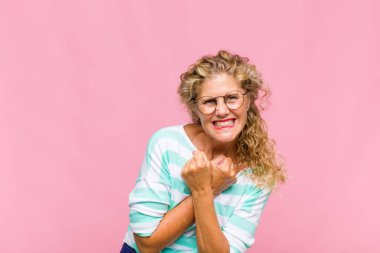 Orta yaşlı bir kadın neşeli bir şekilde gülümsüyor ve yumruklarını sıkmış, kollarını kavuşturmuş, mutlu ve pozitif hissediyor.