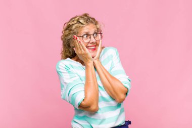 Orta yaşlı bir kadın, mutlu, heyecanlı ve şaşırmış hissediyor, iki eli yüzü yana bakıyor.