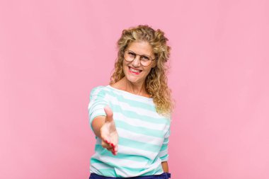 Orta yaşlı bir kadın gülümsüyor, mutlu, kendinden emin ve arkadaş canlısı görünüyor, bir anlaşmaya varmak için el sıkışmayı teklif ediyor, işbirliği yapıyor.