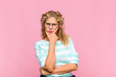 Orta yaşlı bir kadın ciddi, düşünceli ve güvensiz görünüyor, bir kolu çapraz ve çenesi üzerinde, seçenekleri tartıyor.