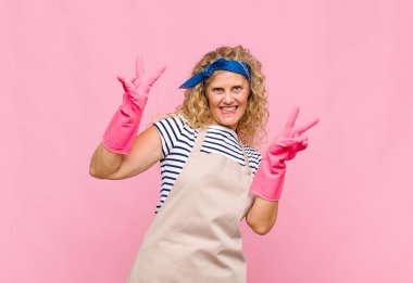 Orta yaşlı kadın gülümsüyor ve mutlu görünüyor, arkadaş canlısı ve memnun, her iki elinle zafer ya da barış işareti yapıyor.