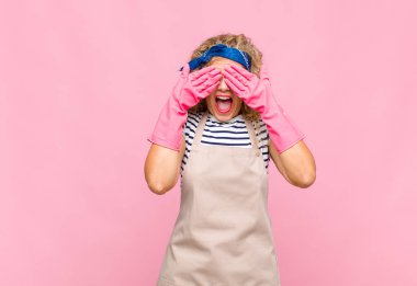 Orta yaşlı bir kadın gülümsüyor ve mutlu hissediyor, iki eliyle gözlerini kapatıyor ve inanılmaz sürpriz ev kadını konseptini bekliyor.