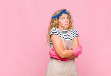 Orta yaşlı bir kadın omuz silkiyor, kafası karışık ve kararsız, kolları bağlı ve şaşkın görünüyor.