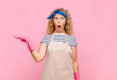 Orta yaşlı kadın şaşırmış ve şoka uğramış görünüyor. Çenesi düşmüş, eli açık bir objeyi elinde tutuyor.