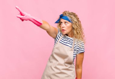 Orta yaşlı bir kadın şaşkın ve şaşırmış hissediyor, hayret içinde, ağzı açık, hayret dolu bakışlı hizmetçi konseptiyle yukarı bakıyor ve işaret ediyor.