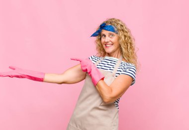 Orta yaşlı bir kadın neşeyle gülümsüyor ve avucunun kenarındaki fotokopi alanını işaret ediyor, nesne temizlikçisi konseptini gösteriyor ya da reklamını yapıyor.