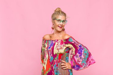 Orta yaşlı bir kadın mutlu, neşeli ve kendinden emin görünüyor. Gururla gülümsüyor ve iki elini de kalçasında istiyor.