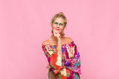 Orta yaşlı kadın mutlu görünüyor ve gülümsüyor. Elleri çenesinde, merak ediyor ya da soru soruyor. Seçenekleri karşılaştırıyor.