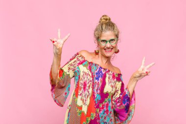 Orta yaşlı kadın gülümsüyor ve mutlu görünüyor, arkadaş canlısı ve memnun, zafer ya da barış işareti yapıyor iki eliyle