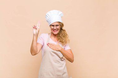 Orta yaşlı kadın fırıncı mutlu, kendinden emin ve güvenilir görünüyor, gülümsüyor ve zafer işareti gösteriyor, olumlu bir tavırla.