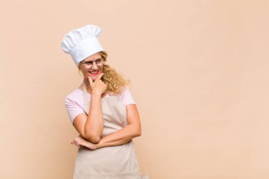 Orta yaşlı fırıncı mutlu, kendinden emin bir ifadeyle gülümseyen, eli çenesinde, merak eden ve yan tarafa bakan.