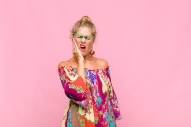 Orta yaşlı bir kadın yanağını tutuyor ve ağrılı diş ağrısı çekiyor. Hasta, sefil ve mutsuz hissediyor. Dişçi arıyor.