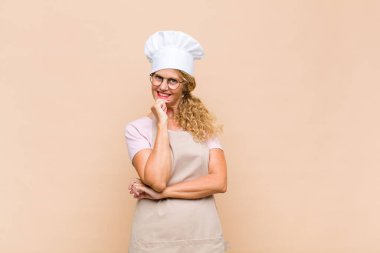 Orta yaşlı fırıncı mutlu görünüyor ve gülümsüyor. Elleri çenesinde, merak ediyor ya da soru soruyor. Seçenekleri karşılaştırıyor.