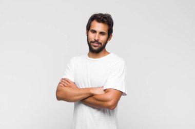 Genç Latin yakışıklı adam kameraya çapraz kollarıyla gülümsüyor ve mutlu, kendinden emin, tatmin olmuş bir yüz ifadesi var.