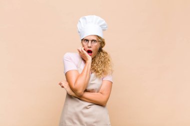 Orta yaşlı kadın fırıncı şok ve inançsızlıkla ağzı açık, elleri yanağında ve kolları çaprazlanmış, sersemlemiş ve şaşkın hissediyordu.