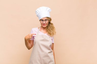 Orta yaşlı kadın fırıncı gururlu, kendinden emin ve mutlu görünüyor, gülümsüyor ve kendini işaret ediyor ya da bir numaralı işareti yapıyor.