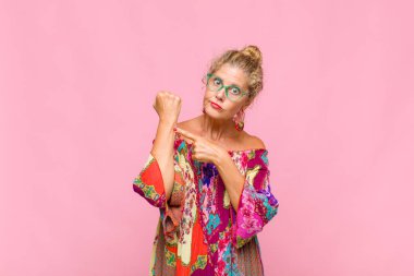 Orta yaşlı bir kadın sabırsız ve kızgın görünüyor, saati gösteriyor, dakiklik istiyor, zamanında gelmek istiyor.