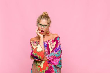 Orta yaşlı bir kadın şok ve inançsızlık içinde, elleri yanağında ve kolları çaprazlanmış, sersemlemiş ve şaşırmış hissediyor.