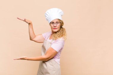 Orta yaşlı fırıncı bir objeyi iki eliyle fotokopi alanında tutuyor, bir objeyi gösteriyor, sunuyor ya da reklamını yapıyor.