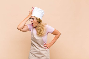 middle age woman baker raising palm to forehead thinking oops, after making a stupid mistake or remembering, feeling dumb