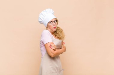 Orta yaşlı fırıncı kadın omuz silkiyor, kafası karışık ve kararsız, kolları bağlı ve şaşkın görünüyor.