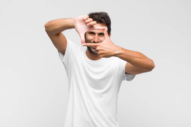 Genç Latin yakışıklı adam mutlu, arkadaş canlısı ve pozitif hissediyor, gülümsüyor ve elleriyle bir portre veya fotoğraf çerçevesi yapıyor.