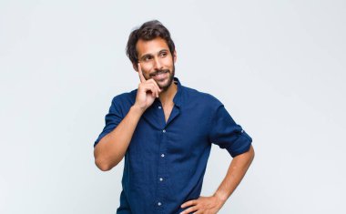 Genç Latin yakışıklı adam mutlu görünüyor ve eli çenesinde gülümsüyor, merak ediyor ya da soru soruyor, seçenekleri karşılaştırıyor.