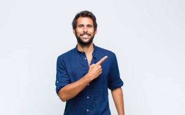 Genç Latin yakışıklı adam neşeyle gülümsüyor, mutlu hissediyor ve kenarı ve yukarıyı işaret ediyor, fotokopi uzayında nesne gösteriyor.